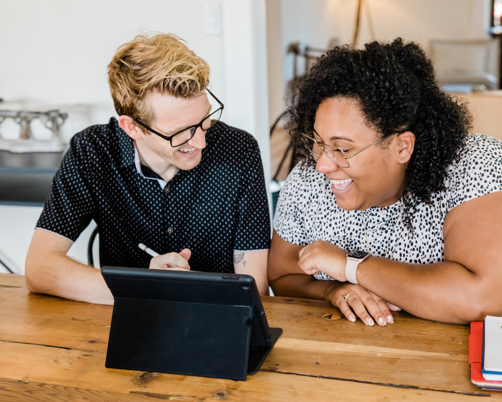A white short haired blond man with spectacles and a curly black woman sit next to each other at a wooden table while looking together towards the screen of a tablet. It looks like they are enjoying their work