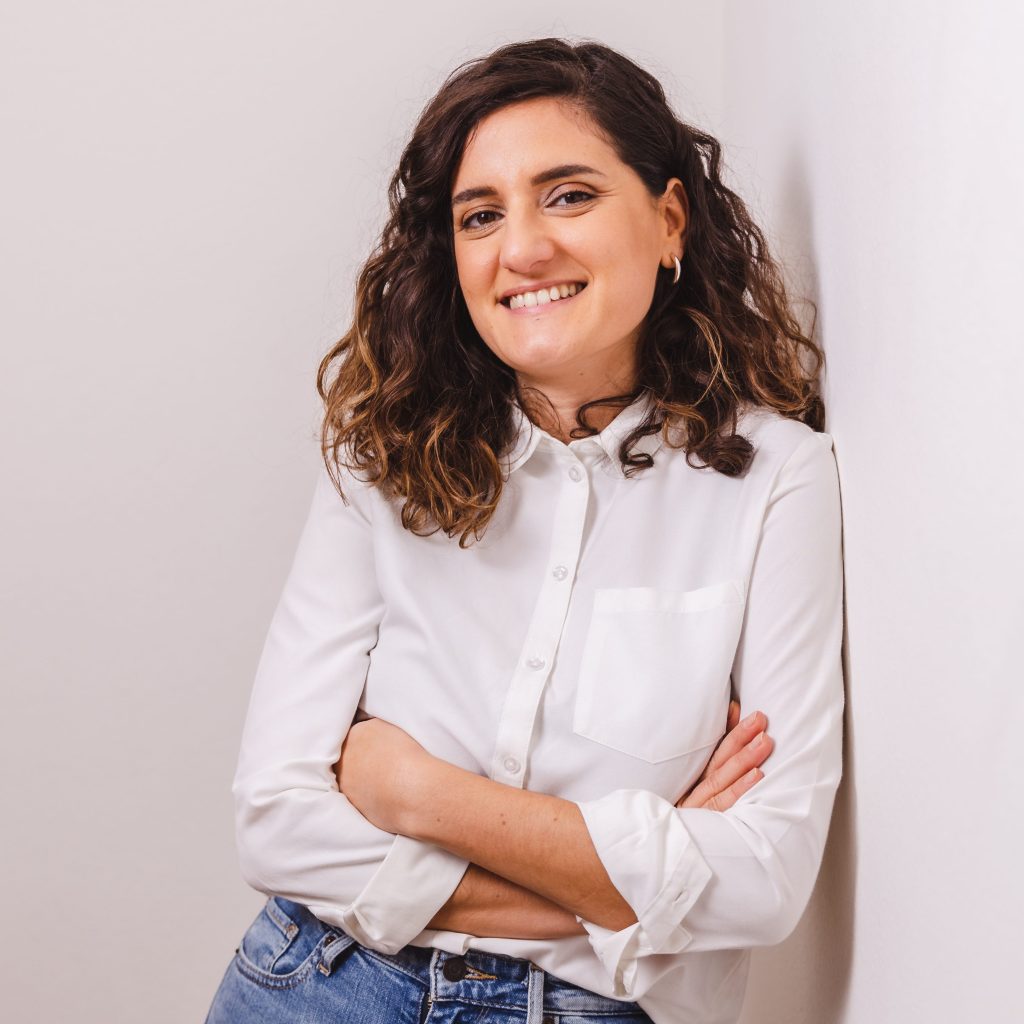 White woman in white shirt, jeans and curly hair leaning towards a wall